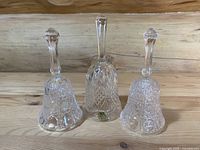 Three clear crystal and cut glass decorative bells arranged side by side on a wood surface, showing variations in glass pattern and shape.