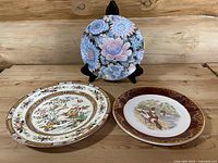 Set of three vintage decorative collector plates shown on wooden surface with stands. Three distinct designs identifiable: floral pastel Toyo plate, maroon bordered Weatherby Royal Falcon plate, and colorful Chinese-style Hong plate.