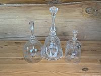 Whole lot of three vintage collectible glass bells displayed on wood surface: Bacalles hand cut starburst, Lord’s Prayer embossed, and bluebird etched bell.