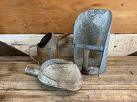 Three vintage galvanized metal farm tools arranged on a wooden surface with a wooden paneled background. Includes a watering can, scoop, and hand shovel.