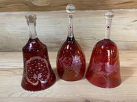 Lot of three ruby red glass bells with clear handles and distinct etched designs on wooden surface.