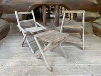 Image showing the set of three weathered wooden folding pieces: two folding chairs and one folding stool/table on a wooden floor in a rustic setting.