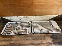 Two white trays with approximately 50 wrenches of mixed sizes and brands, both combination and open-end types arranged on wooden surface.