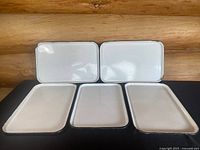 Full view of all five enamelware trays showing white surfaces with black trim edges arranged in two rows on dark surface against wood background