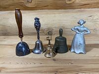 Five vintage metal bells arranged side by side on wooden floor against a wood backdrop, showing full set with mixed styles and finishes.