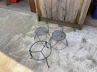 Five wrought iron plant stands grouped on concrete with wooden backdrop. Includes two tall basket style stands, two mesh-top stands, one open base stand.