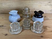 Six vintage insulators arranged on a wooden surface including clear glass, light blue ceramic, and black ceramic styles.