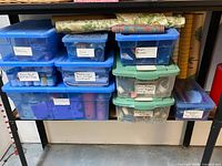 Blue and clear plastic bins stacked and labeled with contents: batteries, cord bundles, snaps and buckles, fabric paint stamps and stencils, patterns for knitting and needlepoint, Velcro ribbing binding and zippers, knitting needles