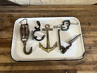 Photo shows six vintage metal hardware items on a white tray including a pulley, anchor hook, two clamps, and two other hooks.