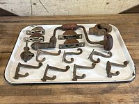 Top-down photo showing vintage cast iron hooks, barn pulleys, hand drill, and miscellaneous hardware on a white tray against a wooden surface.