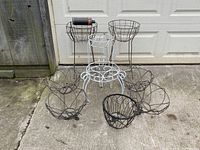 Lot arranged on concrete floor in front of a white garage door showing seven plant holders including wire hanging baskets and stands.