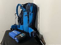 Side view of the MEC Serratus 85L hiking backpack showing blue padded straps and pack design with rain cover and first aid kit placed beside it.