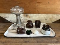Display of all items on a white tray against a wooden background showing clear glass oil lamp base, five ceramic insulators in brown and white, and a green glass ring and furniture foot.