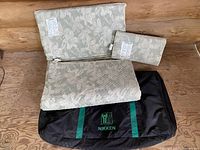 Photo showing the Nikken Kenkopad twin mattress pad, Kenkopillow king size pillow, and Nikken carry bag all together on a wooden floor. The floral patterned fabrics are visible along with the black carry bag featuring green Nikken branding on front.