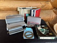 Wide view of all items included in the lot showing the Ibico comb binder, plastic combs box, paper cutter, assorted binders, GEMEX page protectors, metal clips, clipboard, and protractor.