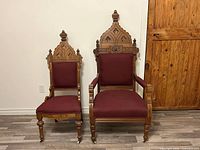 Front view of matched pair of Victorian Gothic ecclesiastical throne chairs showing ornate carved wooden crowns, rich burgundy upholstery, and turned legs with casters.
