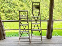 Front view of two weathered wooden ladders of differing heights on porch, showing rustic wear and crossbeam supports.