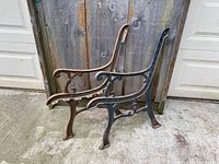 Pair of vintage cast iron garden bench ends, one showing rusty brown patina, the other with black paint traces, placed on concrete against a wooden fence.