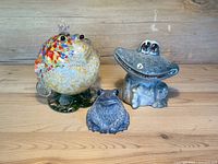Front and side views of the three frog figurines grouped together on wooden surface against rustic wood background. The colorful glass frog with crown, blue-green ceramic wide-mouth frog, and small gray soapstone frog shown.