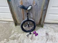 Photo showing the Kris Holm trials unicycle upright against wooden fence with purple diabolo, wooden sticks, and metal clown horn on the ground beside it.