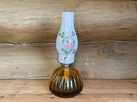 Oil lamp standing assembled with amber ribbed glass base and white glass chimney painted with pink flowers and green leaves.