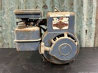 Side view of the vintage Briggs & Stratton engine showing the blue painted body, recoil starter, fuel tank, and decal plate.