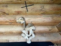 Plush camel marionette puppet hanging from a black wooden crossbar controlled by strings. Shows full puppet body and face details in front of wood log wall.