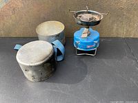 Full view of vintage Camping Gaz Globe Trotter stove with metal cooking pot set and storage case on dark surface, showing blue stove and silver metal pots with blue handles.