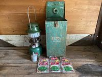 Photo showing the Coleman lantern, original green metal carrying case, metal fuel funnel, and three packs of sealed mantles displayed together on a wooden surface.