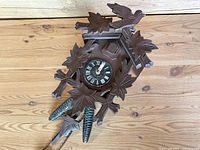 Front view of vintage German cuckoo clock with carved wood leaves and bird, pinecone weights dangling below.