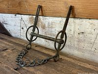 Photo of Victorian era handcrafted solid iron horse bridle bit with attached iron chain, showing patina and aged condition on wooden surface.