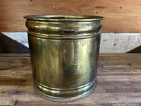 Front and side view of the large cylindrical brass planter showing its polished patina and ring decorations near the rim and base, placed on wooden surface.