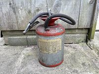 Full view of the vintage cylindrical metal oil sprayer can with red top and bottom accents, attached black rubber hose with trigger nozzle. Shows wear and rust spots on body and hose.