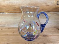 Full side view of the clear glass Murano pitcher with colorful millefiori floral patterns and cobalt blue handle and base on wooden surface.