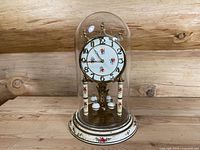 Full view of vintage Kundo clock under glass dome showing floral design and brass base on wooden surface with log wall background