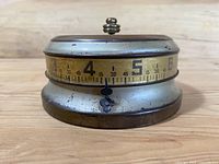 Side view of the clock showing the rotating brass dial with numbers 4 and 5 visible, and the fixed black pointer at the base.