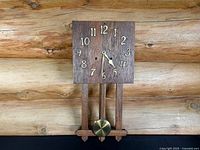 Full view of the wooden mission-style wall clock with square face, brass numerals and brass pendulum on an elongated wooden frame against a log wall.