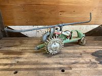 Full side view of vintage walking sprinkler showing spiked metal wheels and long handle laid on wooden surface.