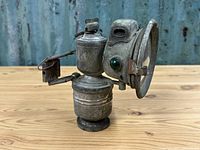 Side view of vintage metal carbide lamp showing green glass side jewel, mounting bracket, and fuel tank behind main lamp housing