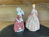 Two vintage porcelain lady figurines, one in pink gown with floral designs and the other in darker fur-trimmed dress, standing on wooden surface.
