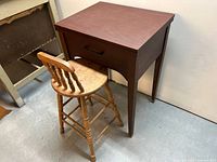 Front angled view of vintage sewing desk with single drawer, showing fold-out top and dark wood finish, accompanied by light wood spindle-back stool with turned legs.