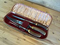 Open fitted case with red velvet and satin lining showing carving knife, fork, and sharpening steel with antler handles.