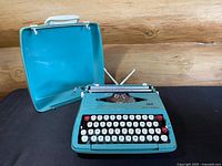 Teal blue Smith-Corona Corsair typewriter with matching open hard-shell case, showing front and side views.
