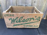 Front view of wooden crate showing 'Drink Wilson’s Since 1875 Ginger Ale' in faded green lettering with visible wear and wood grain.