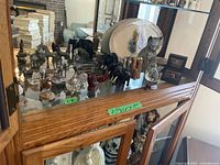 Display cabinet top shelf with multiple metal and wooden animal figurines, metal trinket box, and serving platter with crazing.