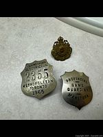 Three badges on a light-colored surface: Toronto Journeyman Electrician badge, Manitoba Provincial Game Warden badge, Royal North West Mounted Police badge.