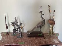 Photo showing the full lot on a marble surface including the teak crane sculpture, metal peacock sculpture on stand, metal tree sculpture holding birds and flowers, and the matching decorative candle holders.