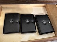 Photo shows three black leather-like coin cases with a silver snap button and an embossed maple leaf, all unopened, arranged side by side in a drawer.