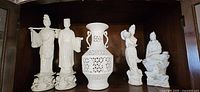 Front view of five white porcelain Chinese figurines and one pierced lattice vase displayed on a dark wood shelf.