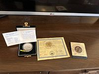 Display of three collectible coins and their certificates: Chinese silver 5 Yuan coin in black case, U.S. $5 gold proof coin with certificate, Masada tribute token with its certificate.